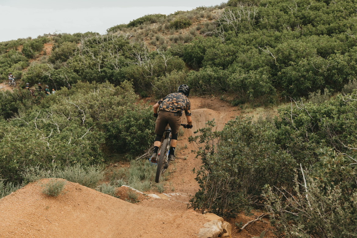 A mountain biker dressed in a dark, patterned shirt and brown shorts is seen jumping off a dirt ramp on a trail surrounded by green shrubs and trees. The rider is captured mid-air, with a sloped hillside in the background and several other cyclists visible in the distance.