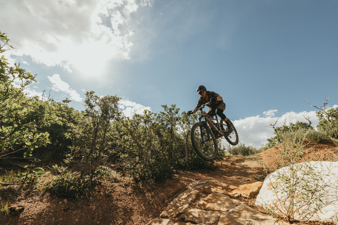 A mountain biker jumps off a dirt path, surrounded by greenery and under a bright blue sky. Sunlight shines above, casting a dramatic light on the scene. The rider is wearing a helmet and a patterned jersey. The terrain features rocky sections and shrubs, indicating a natural outdoor biking trail.