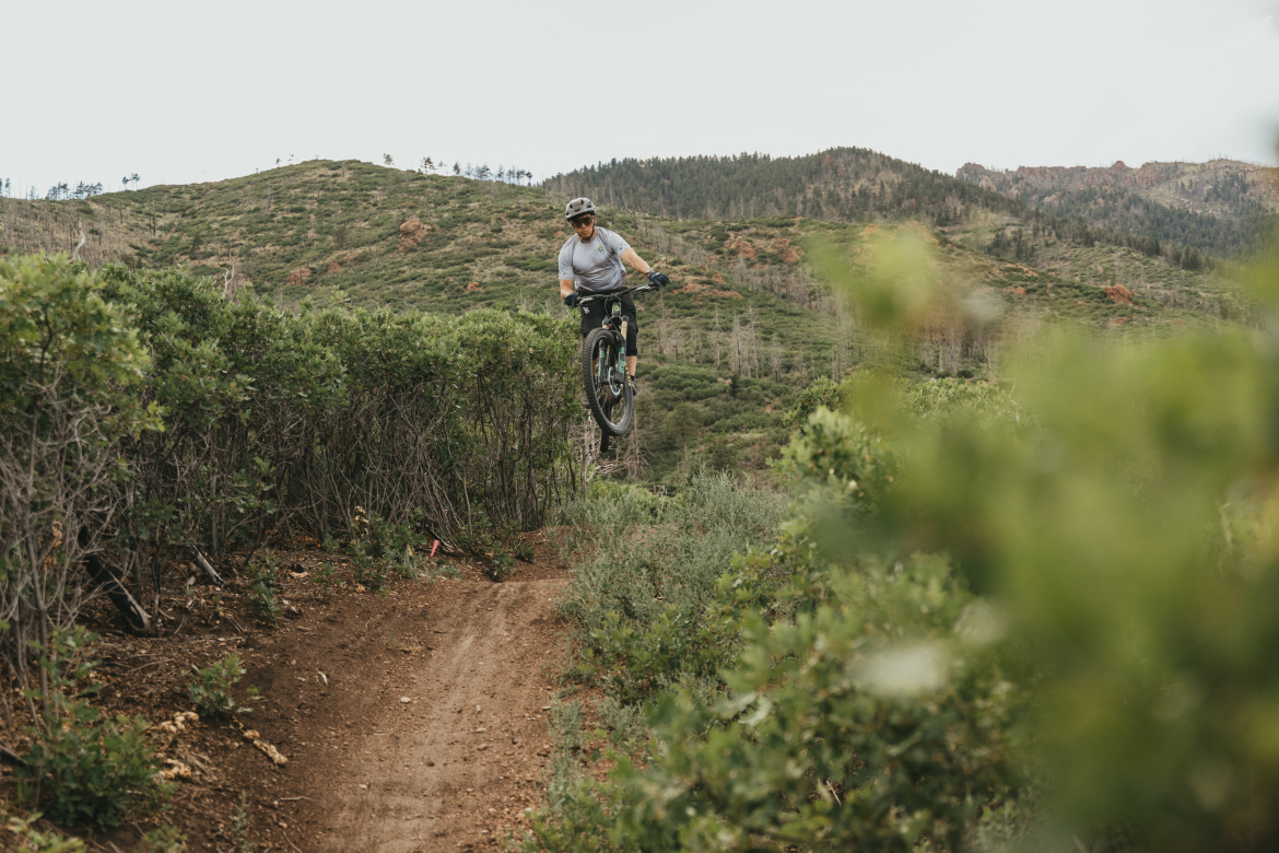 A mountain biker performing a jump over a dirt trail surrounded by shrubs, with rolling green hills and trees in the background. The sky is slightly overcast, creating a natural outdoor setting for the activity.