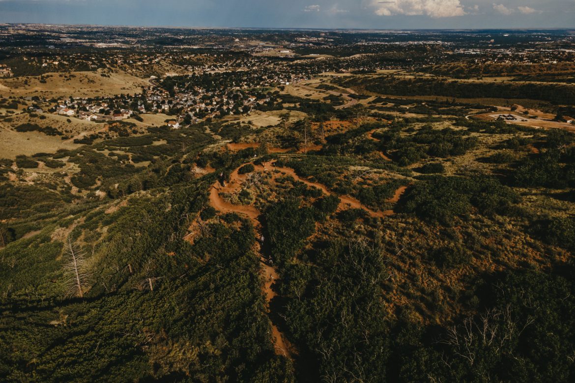 Aerial view of a hilly landscape featuring green vegetation and reddish earth, with a small residential area visible in the background. The scene showcases winding trails through the terrain, along with patches of trees and open fields, under a clear sky with a few clouds.