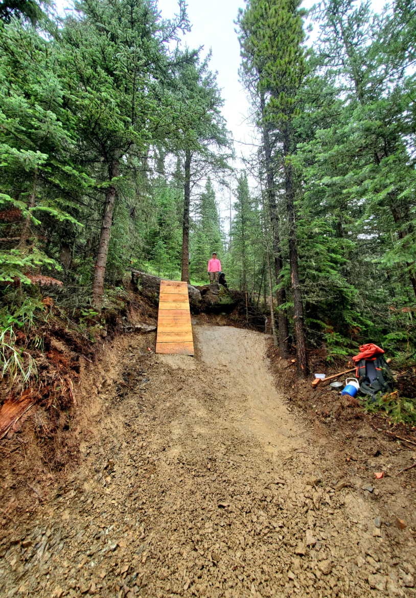 A mountain biker standing at the top of a wooden ramp on a dirt trail, surrounded by tall green trees in a forested area. The trail is slightly muddy, and there are tools and clothing on the ground nearby.