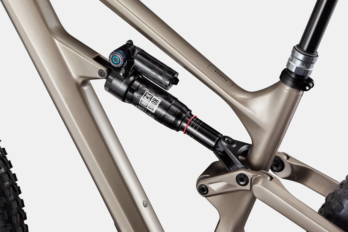 Close-up view of the suspension system on a mountain bike frame, featuring a RockShox shock absorber mounted on a sleek, bronze-colored frame. The image highlights the intricate design and engineering of the bike's components.