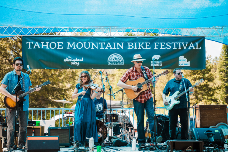 A live band performing on stage at the Tahoe Mountain Bike Festival, with a banner displaying the festival name in the background. The musicians include a guitarist, a female violinist, and a male guitarist, all dressed casually and engaging with the audience. The stage is set outdoors under a blue canopy, surrounded by trees.