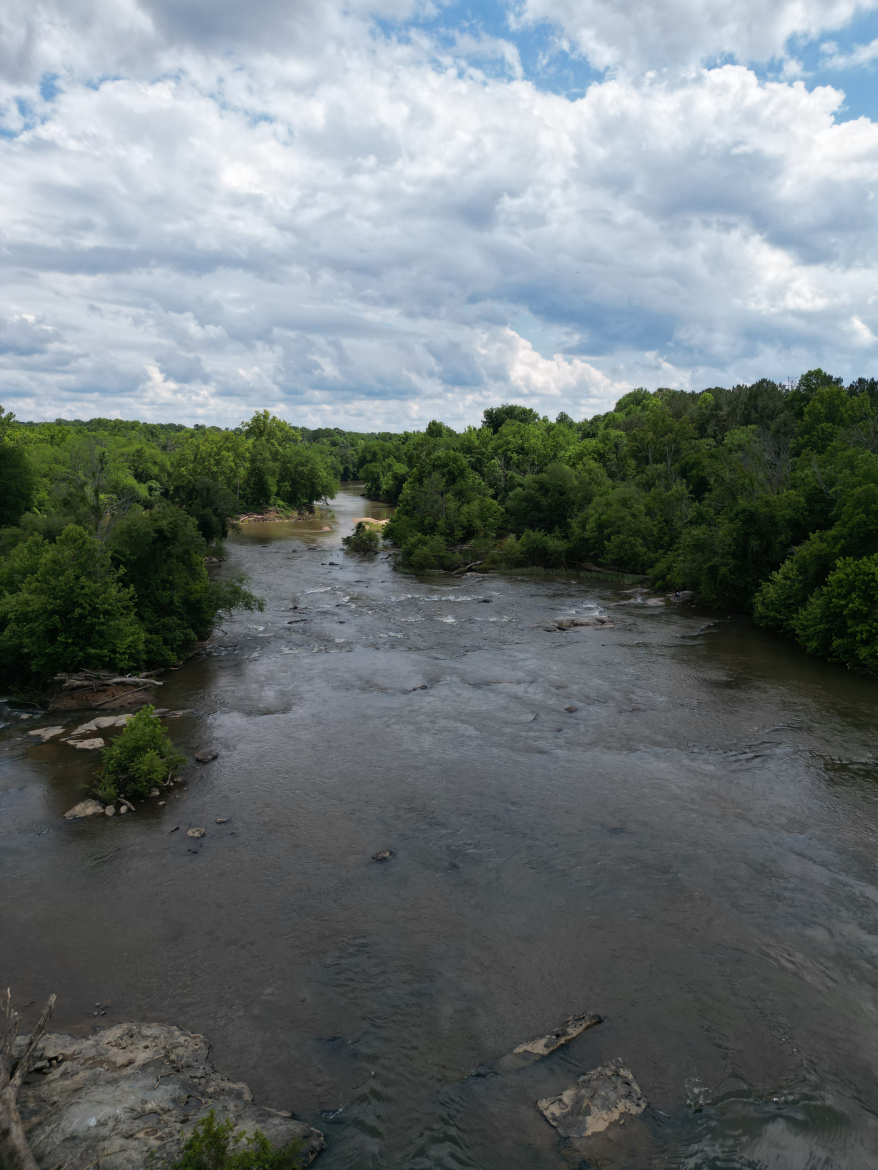 A scenic view of a winding river surrounded by lush greenery under a partly cloudy sky. The water reflects the clouds, and rocky outcrops are visible along the riverbanks.