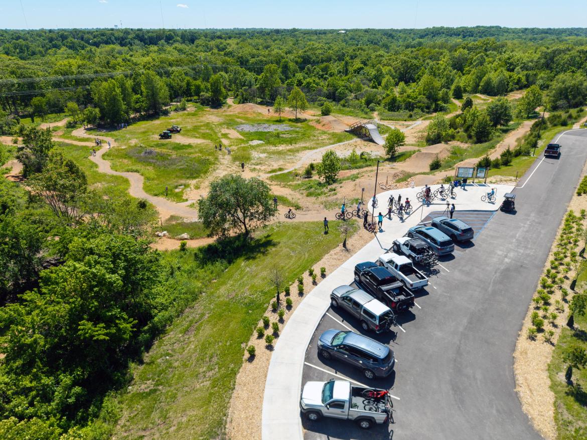 Aerial view of a bike park featuring a parking area filled with vehicles, surrounded by lush greenery. Bicyclists are gathering near the trailhead, while dirt paths weave through the park's grassy landscape, leading to various biking features in the background.