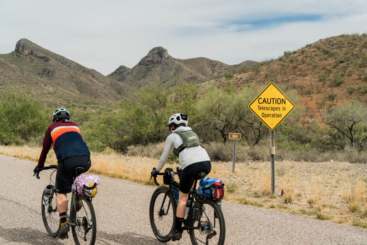 Two cyclists ride along a gravel road surrounded by mountains. A yellow caution sign indicating "Telescopes in Operation" is visible alongside the road, with a mile marker sign labeled "184." The landscape features dry grass and sparse vegetation under a partly cloudy sky.