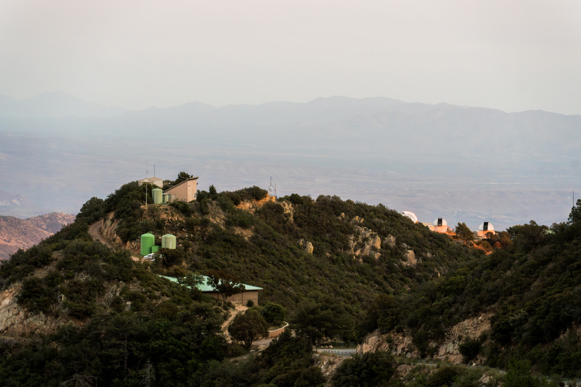 A scenic view of a mountainous landscape featuring several structures atop a hill. The foreground shows a mix of greenery and rocky terrain, with two green water tanks visible. In the background, more buildings and observatories are perched on the hillside, set against a hazy sky and distant mountains.