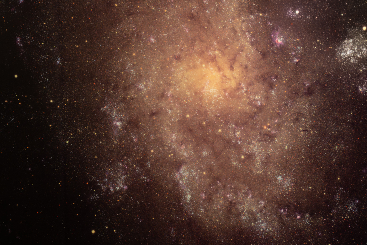 A vibrant view of a spiral galaxy, featuring a swirl of stars, dust, and cosmic formations against a dark background. Shades of orange, purple, and white illuminate various regions, highlighting the galaxy's structure and the density of stars.