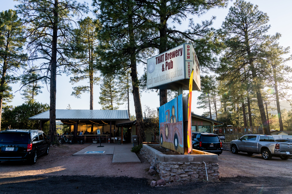 A brewery and pub named "THAT Brewery & Pub" is seen surrounded by tall pine trees. The establishment features a sign indicating it is open and serves locally made beers and food. A colorful mural of a vintage truck is displayed next to the entrance, and several vehicles are parked nearby. The scene captures a relaxed outdoor atmosphere.