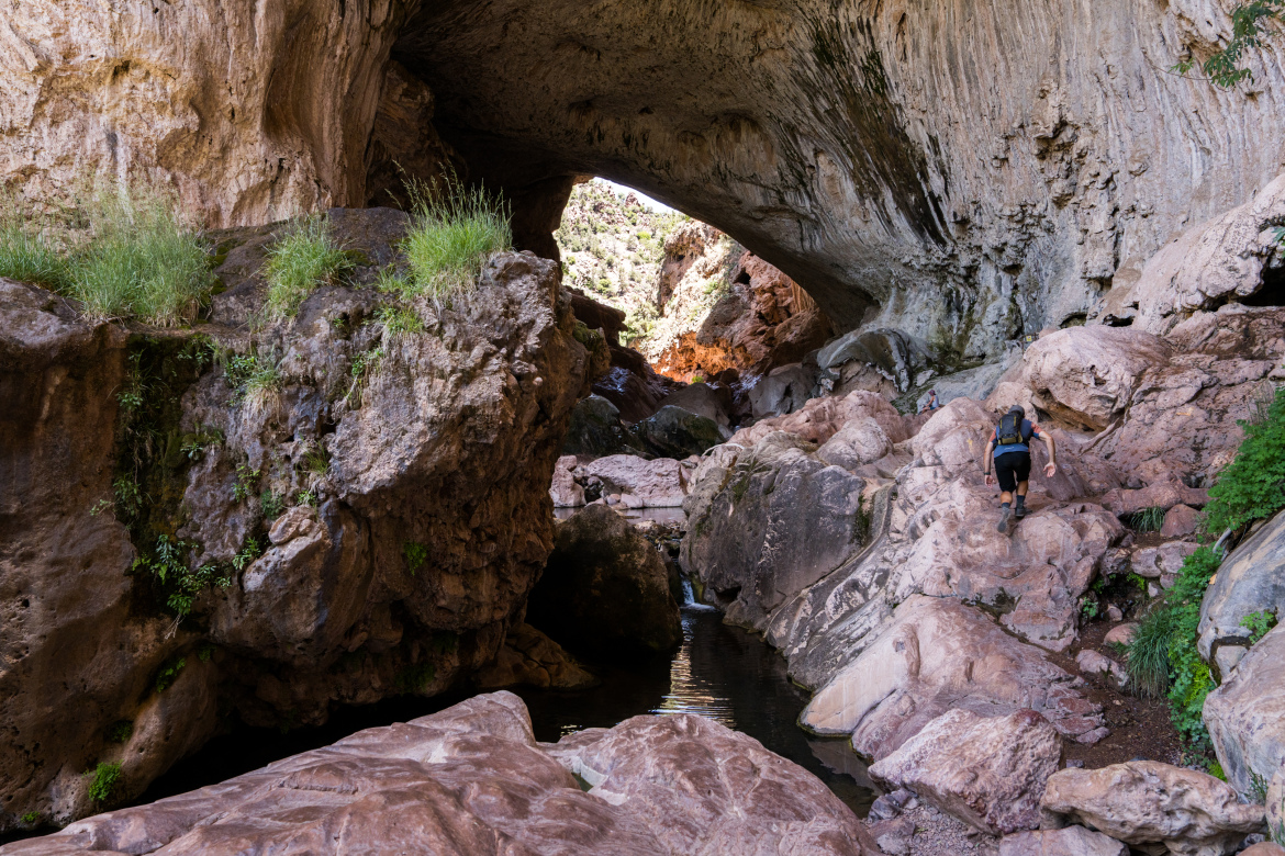 A hiker ascending rocky terrain beneath a large natural rock arch, surrounded by towering cliffs and patches of greenery, with a shallow pool of water visible among the rocks. The scene captures the beauty of a canyon environment.