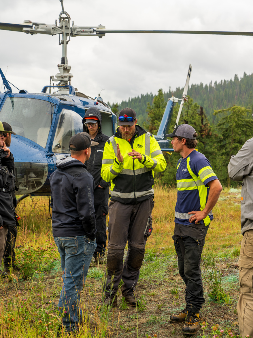 A group of individuals wearing outdoor work clothing are engaged in a discussion in front of a blue helicopter, set amidst a grassy area with trees in the background. Some participants are listening attentively, while one person gestures as they speak. The sky is cloudy, suggesting overcast weather conditions.