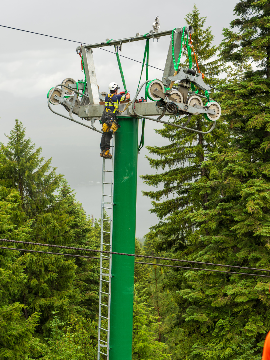 A technician wearing a helmet and climbing gear works near the top of a green ski lift tower surrounded by dense evergreen trees. The technician is adjusting equipment on the lift's drive mechanism, with one foot on a ladder and tools in hand. The sky is overcast, indicating a possibly rainy day.