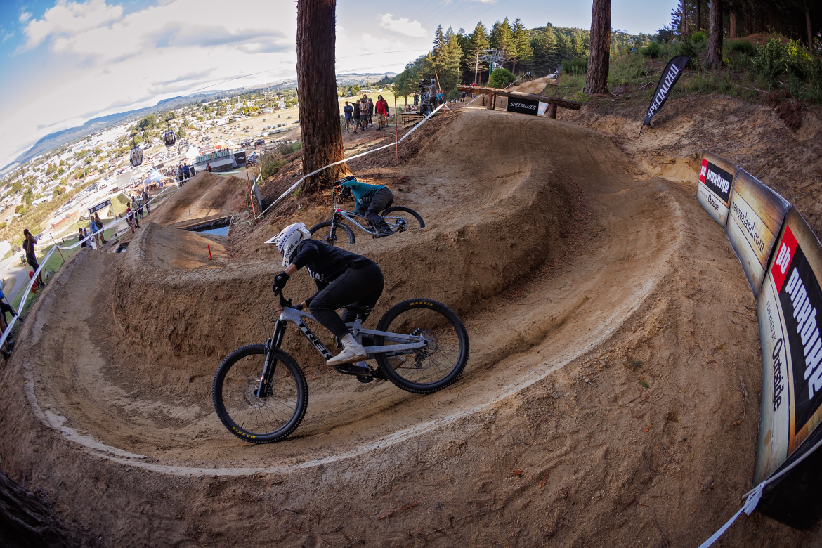 Two mountain bikers navigating a dirt track with curved paths, surrounded by trees and an audience in the background. The setting appears to be a mountain biking event, showcasing a scenic view of the landscape and additional spectators watching the riders.