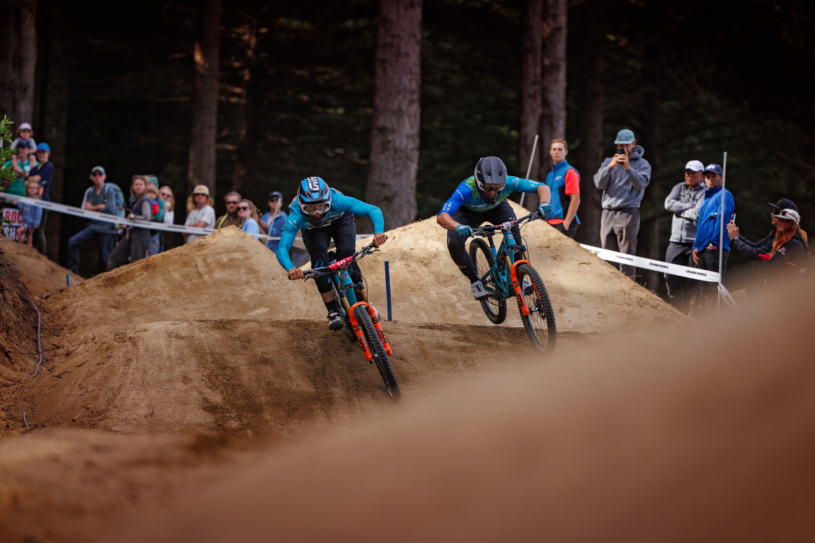 Two mountain bikers in action, racing on a dirt track with jumps in the background. Spectators watching the event are visible in the background among tall trees. The riders are dressed in blue and black gear, and the scene captures the excitement of the competition.