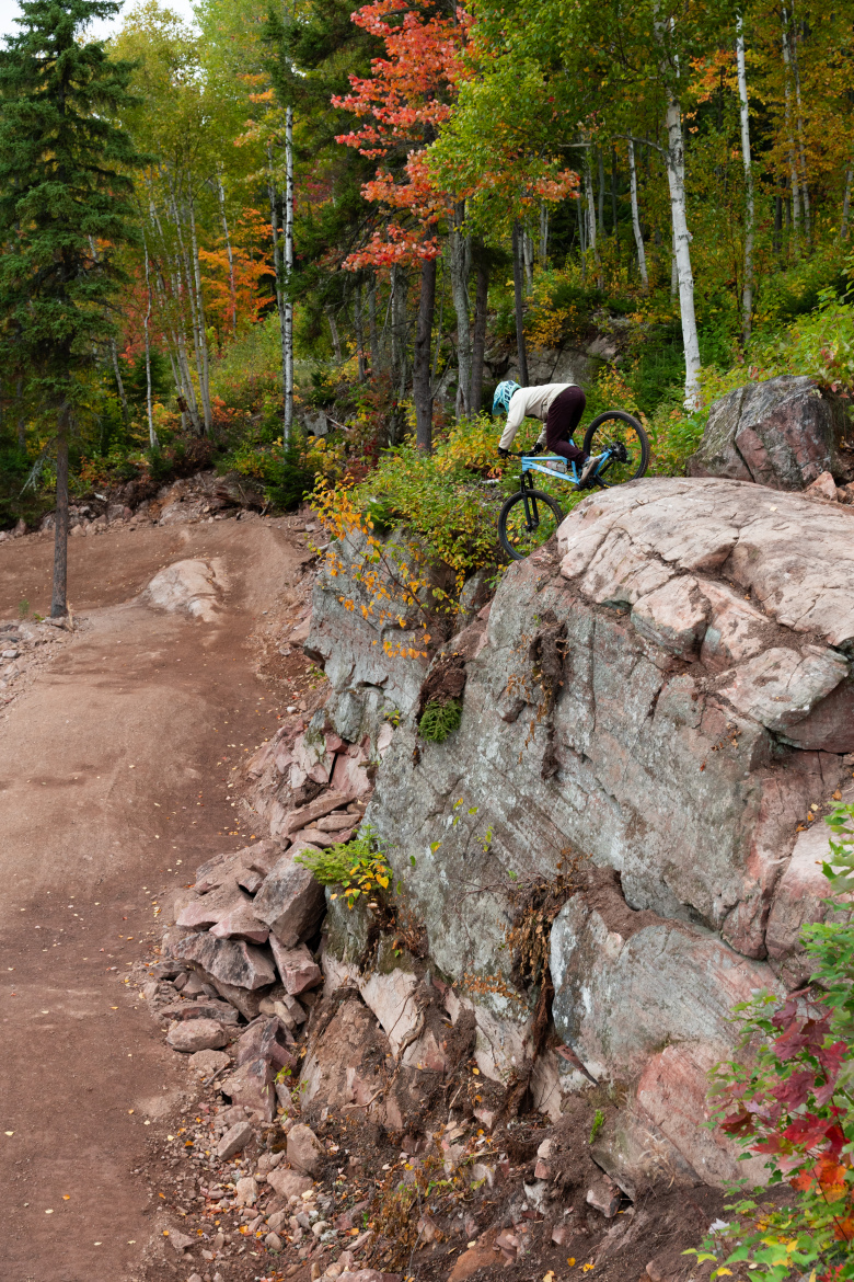 A mountain biker poised on a rocky ledge, preparing to descend a steep trail. Surrounding the biker are vibrant autumn foliage and towering trees, creating a picturesque natural backdrop. The terrain features a mix of dirt paths and rocky outcrops, emphasizing the adventurous setting.
