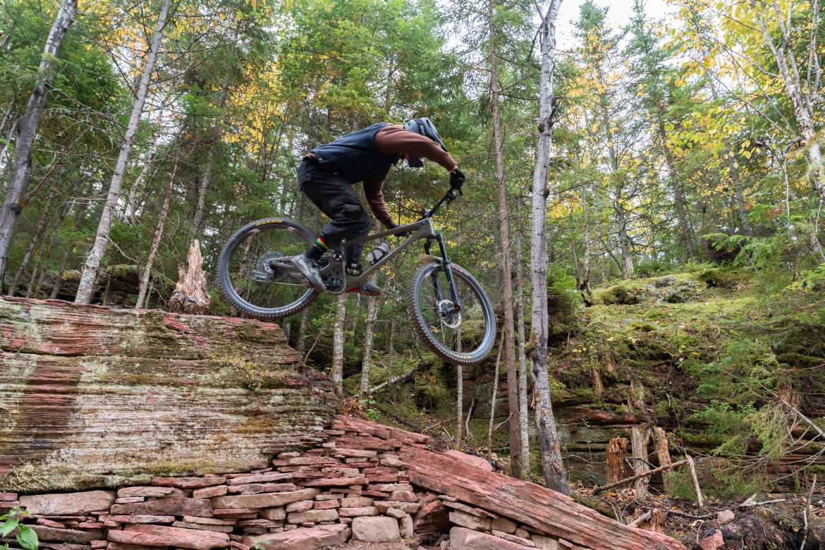 A mountain biker in mid-air above a rocky outcrop in a forested area, surrounded by tall trees and autumn foliage. The biker is wearing protective gear and appears to be executing a jump while riding a mountain bike.