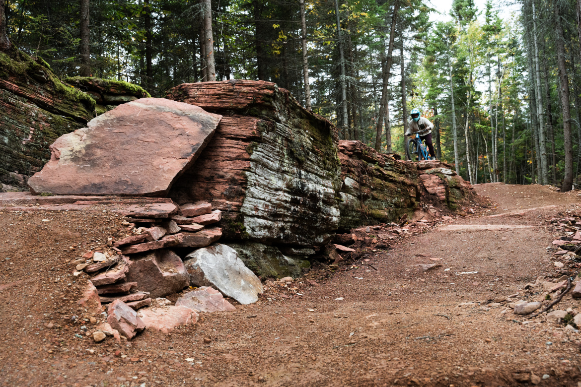 A mountain biker navigates a rugged trail featuring large rock formations and earthy textures, surrounded by tall trees in a forested setting. The path is winding, with layers of reddish-brown rock visible along the side, indicating a natural landscape ideal for off-road biking.