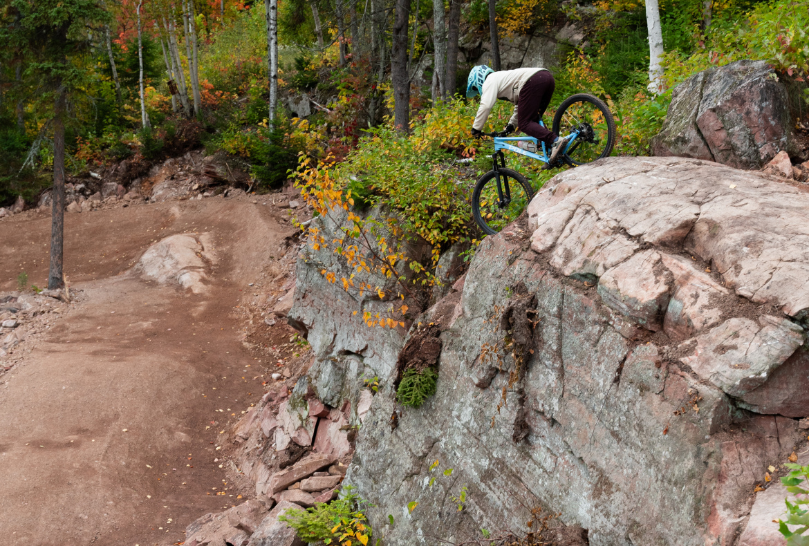 A mountain biker navigates a rocky ledge in an autumn forest, balanced on their bike as they prepare to descend. Vibrant fall foliage adds color to the scene, with trees in the background and a dirt trail visible below the rocks.