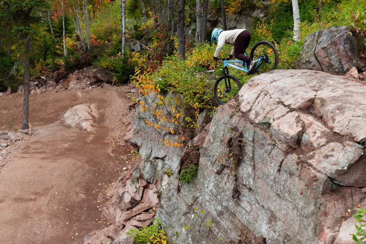 A mountain biker navigates a rocky ledge in an autumn forest, balanced on their bike as they prepare to descend. Vibrant fall foliage adds color to the scene, with trees in the background and a dirt trail visible below the rocks.