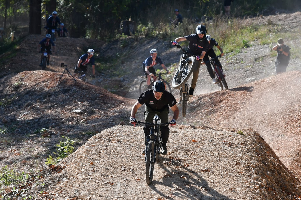 A group of mountain bikers performing tricks on a dirt course in a wooded area. One biker is in mid-air executing a jump, while another is climbing uphill on a bike. Dust is kicked up around the riders, and several spectators can be seen in the background.