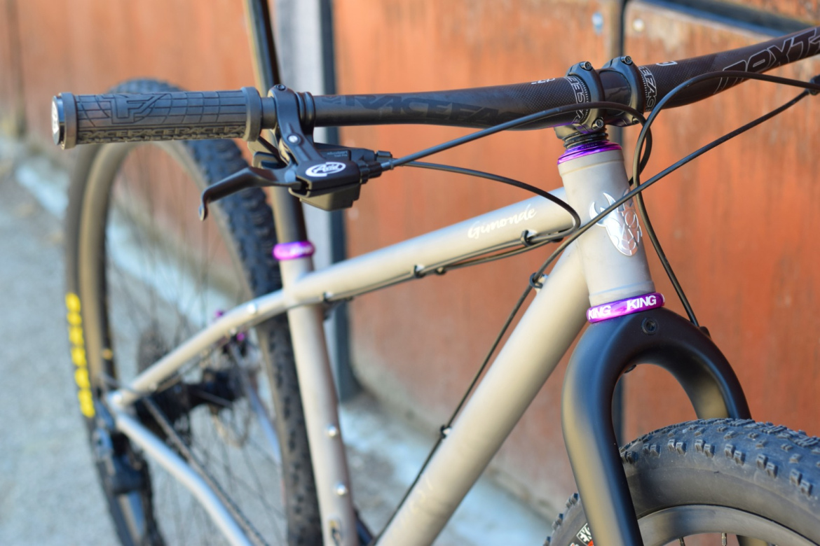 Close-up image of a modern mountain bike highlighting the handlebars, grips, and front fork. The bike features a sleek, silver frame with purple accents, and the background shows a textured orange wall. The focus is on the bike's components, showcasing its design and detailing.