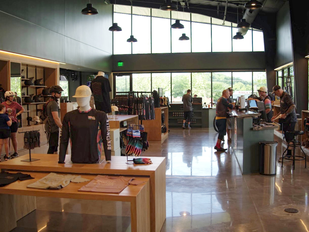Interior of a modern retail store featuring biking gear, with displays of clothing and accessories. Several customers are browsing, and employees are at the checkout area. Large windows allow natural light to fill the space, highlighting the contemporary design and products available.