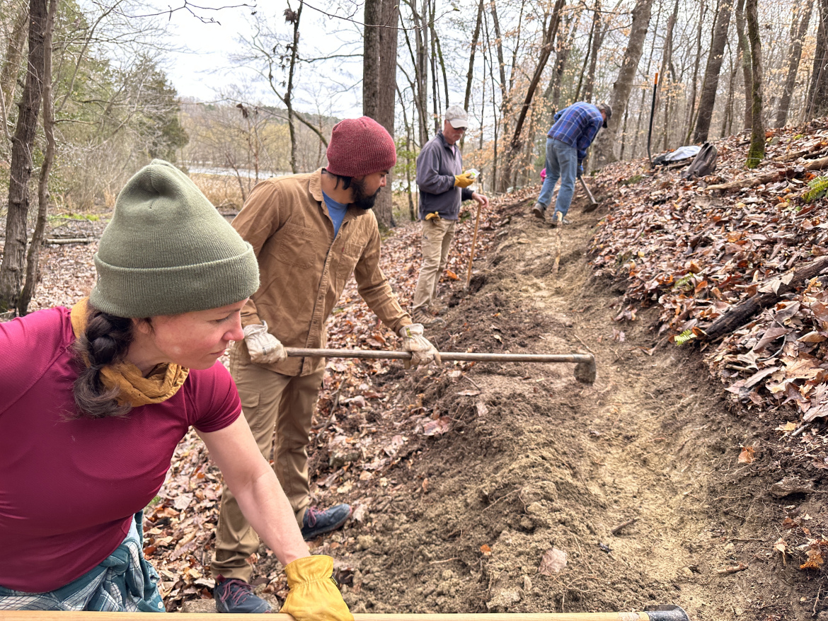 A group of four individuals is working together to maintain a hiking trail in a wooded area. Two people are visible in the foreground, one using a hoe and the other raking soil. Both are wearing gloves and casual outdoor clothing, with a focus on a woman in a green beanie and purple shirt. In the background, two other individuals are digging further up the trail, surrounded by fallen leaves and trees. The scene captures a collaborative effort in an outdoor setting during a cool day.
