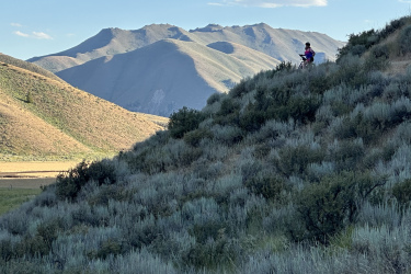 A person standing on a hillside covered with green and brown shrubs, overlooking a valley with distant mountains under a blue sky with a few clouds. Quigley Loop mountain bike trail.