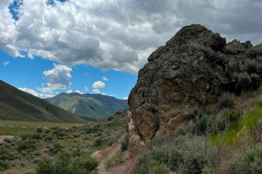 A dirt path winds through a mountainous landscape, flanked by a large rocky outcrop on the right and patches of greenery on either side. The sky is partly cloudy with vibrant blue patches peeking through. Soft hills rise in the background, showcasing the natural beauty of the area. Quigley Loop mountain bike trail.