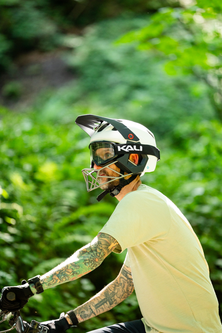 A person wearing a white helmet and protective goggles, with visible tattoos on their arms, rides a bike through a lush green environment. The sunlight filters through the trees, creating a vibrant backdrop.