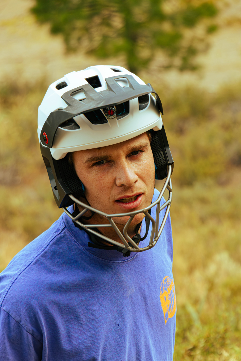 A young man wearing a white and black sports helmet, with a gray mesh faceguard, looks at the camera with a serious expression. He is dressed in a blue T-shirt and is surrounded by a natural landscape with soft earth tones and blurred greenery in the background.