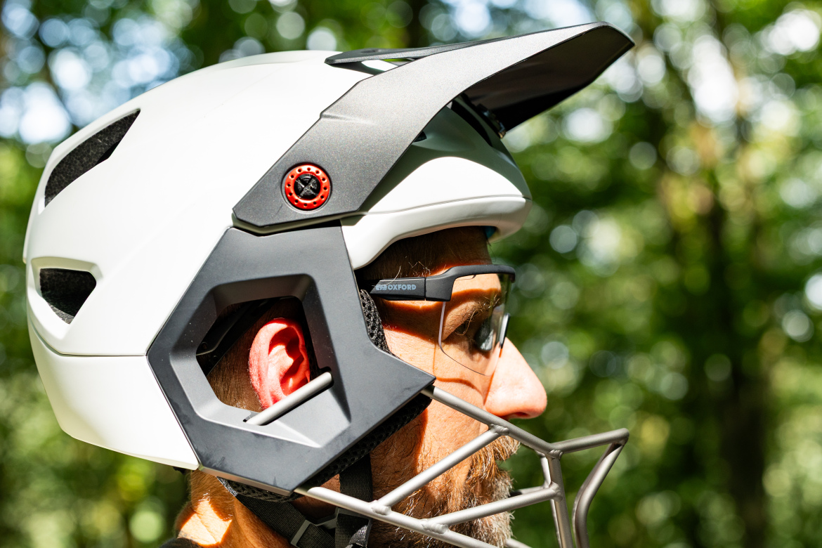 A close-up profile view of a person wearing a white and gray mountain biking helmet, equipped with a peak and ventilation openings. The individual is also wearing clear protective eyewear, set against a blurred green background of trees, suggesting an outdoor environment.