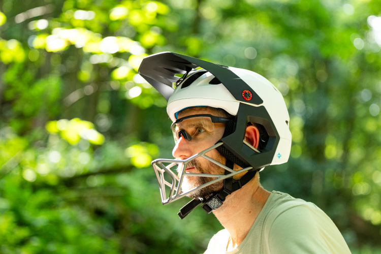 A man wearing a white and black mountain biking helmet and protective eyewear, looking to the side in a forested area with sunlight filtering through the trees.
