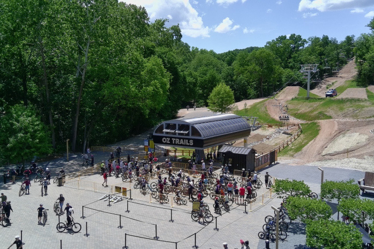 A view of the Oz Trails bike park on a sunny day, featuring a crowd of mountain bikers gathered near the lift station. The area is surrounded by lush greenery and includes bike trails visible in the background. Several bicycles are parked, and people are waiting in line to access the trails.