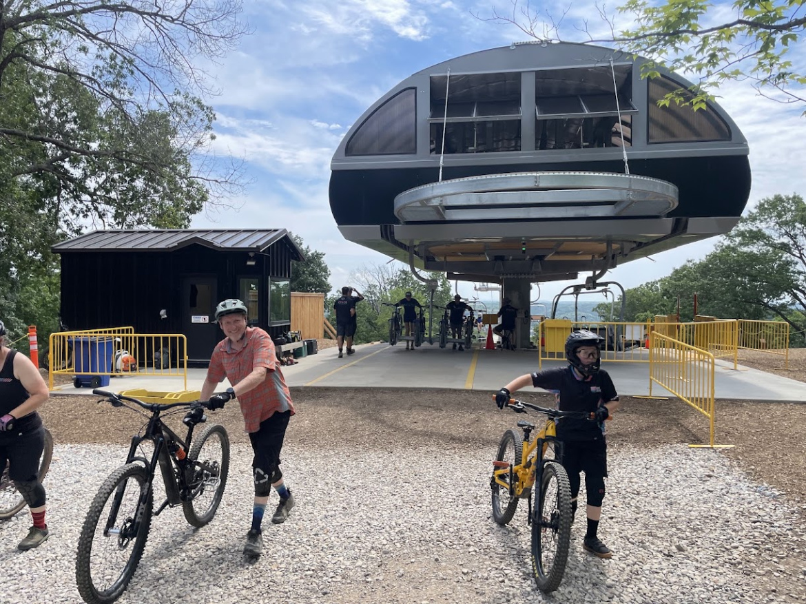 A group of mountain bikers, including two carrying their bikes, walk towards a bike lift station under a cloudy sky. In the background, a large gondola lift is visible above, with a small building nearby. Various people are seen getting ready to ride, surrounded by trees and a gravel path.