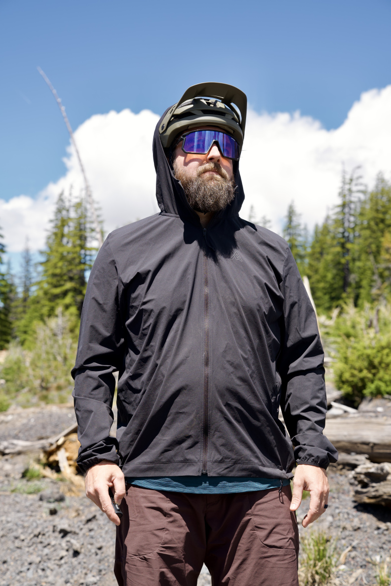 A man with a beard stands outdoors, wearing a black zip-up jacket with the hood up. He has on a green bike helmet and mirrored sunglasses, and is surrounded by a forest with blue sky and clouds in the background.