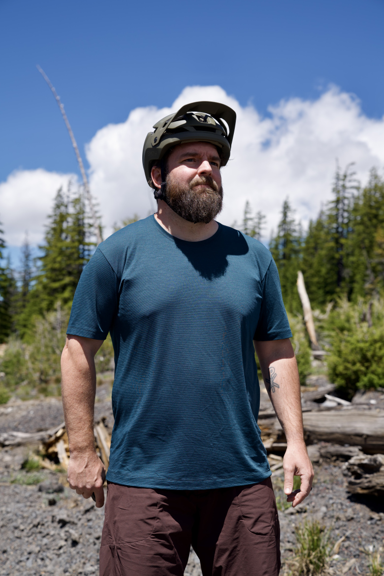 A man with a beard wearing a blue athletic shirt and a dark helmet stands outdoors, surrounded by tall green trees and a clear blue sky with a few clouds. The rugged landscape features scattered rocks and fallen logs.