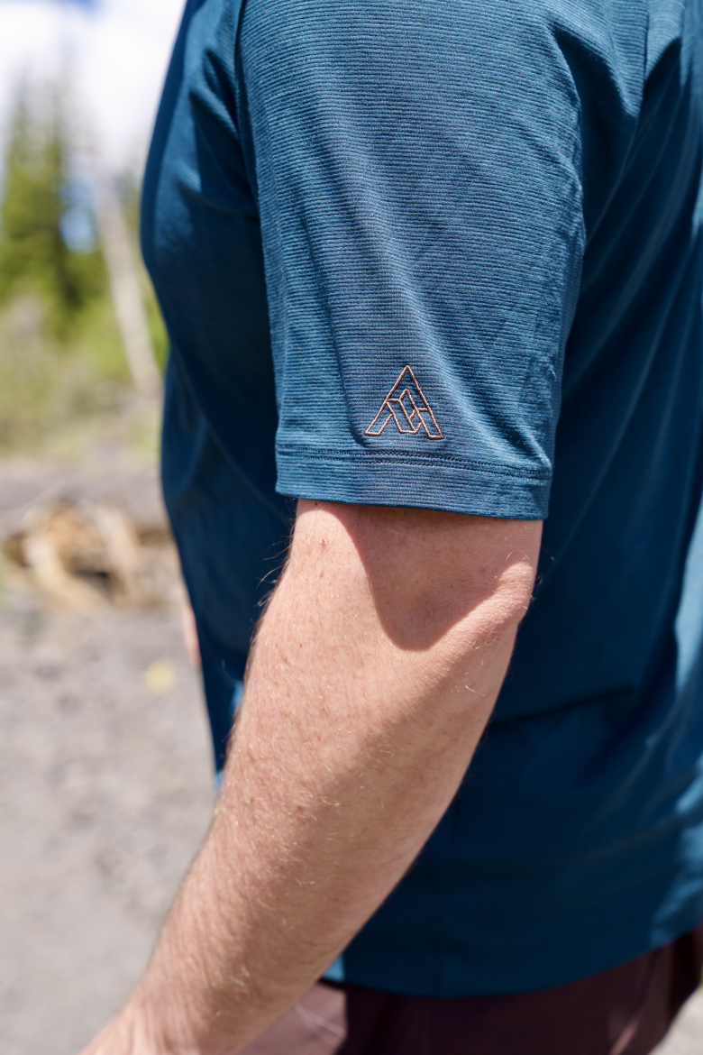 A close-up image of a person's arm wearing a blue athletic shirt with a textured pattern, featuring a small stylized logo on the left sleeve. The background shows a natural outdoor setting with trees and a clear sky.