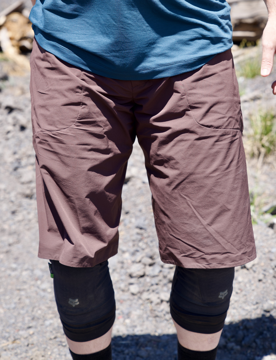 A person is standing outdoors wearing a blue short-sleeve shirt and brown shorts, with leg sleeves that reach below the knee. The background features a rocky terrain with some greenery and wood logs.