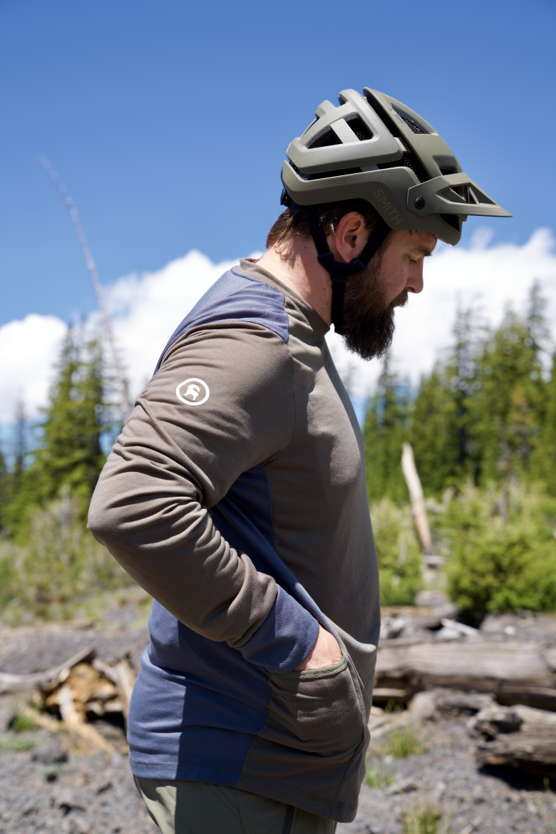 A person wearing a gray and blue long-sleeve shirt and a green helmet stands outdoors, looking to the side. They have a beard and are in a natural setting with trees and a blue sky. Their hands are tucked into the pockets of their shirt.