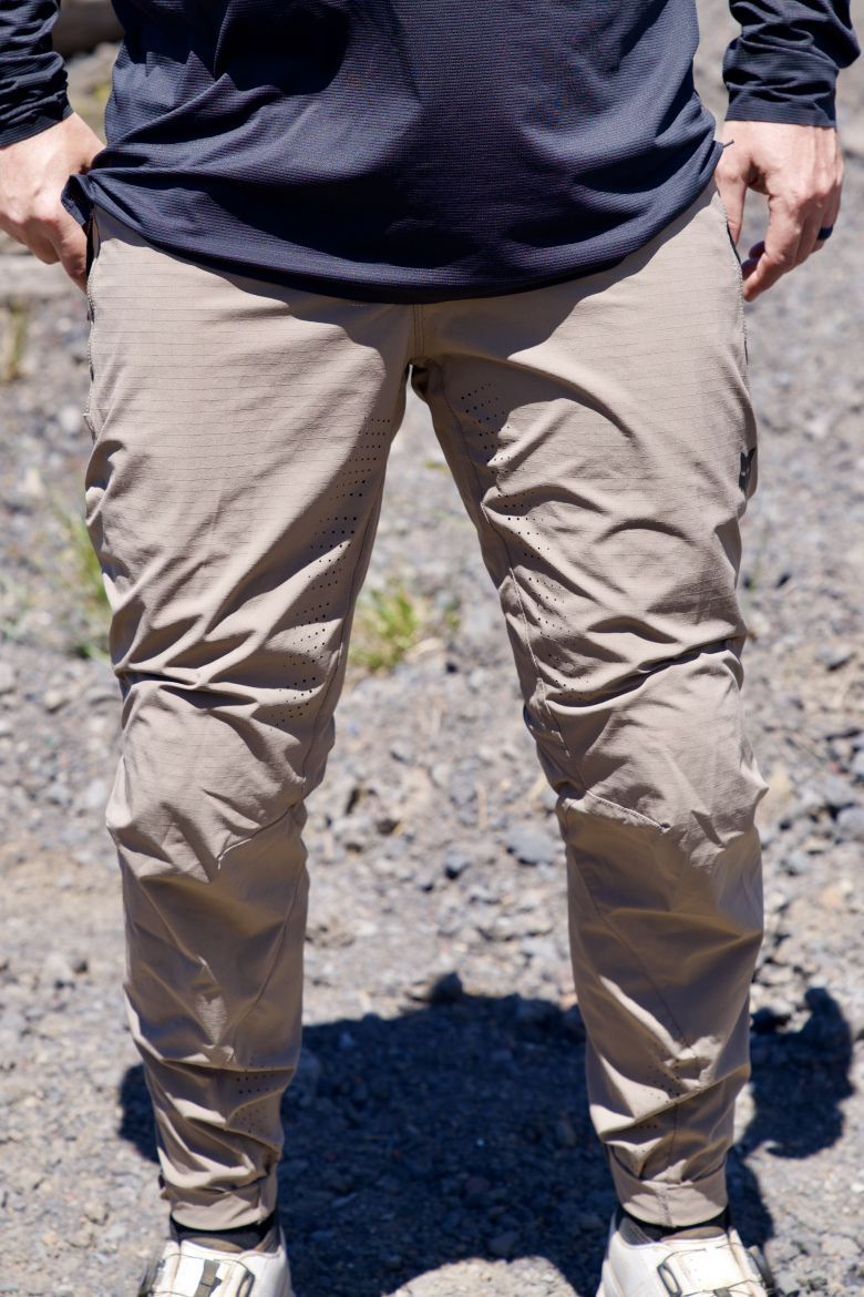 A person stands outdoors wearing light brown pants and a dark long-sleeve shirt, with their hands resting at their sides. The background features rocky terrain and sparse greenery.