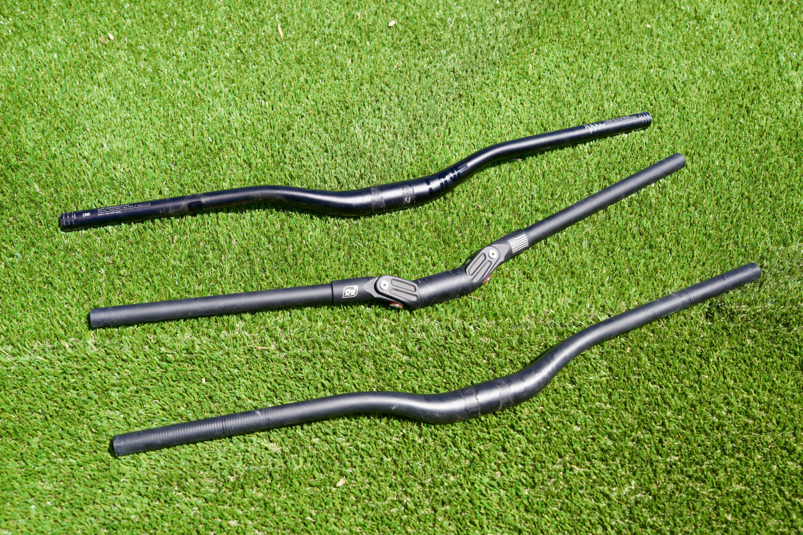 Three bicycle handlebars are displayed on green artificial grass. The top handlebar has a slight curve, while the middle one features a joint and an adjustment mechanism. The bottom handlebar is straight with grip patterns along the sides.