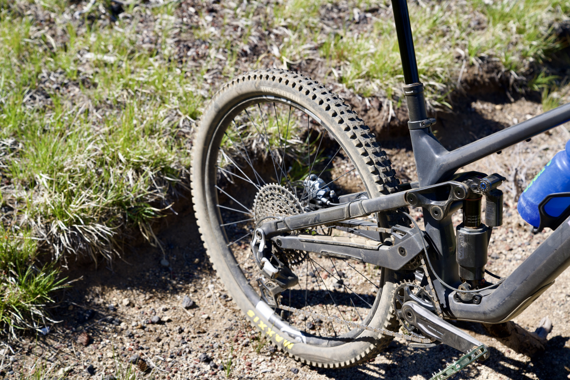 Close-up view of the rear wheel and suspension system of a mountain bike, set against a backdrop of dirt and grass. The wheel features a knobby tire, and the bike frame is partially visible, showcasing its robust design and gearing system.