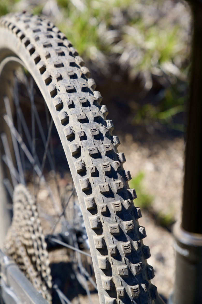 A close-up view of a bicycle tire with a heavily treaded surface, showing dirt and wear. The background is blurred, featuring grass and natural terrain.