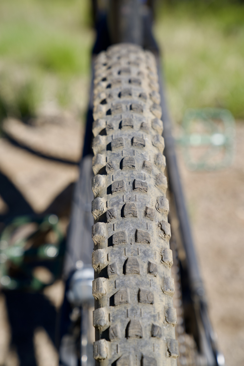 Close-up view of a bicycle tire with a textured tread pattern, showing dirt and wear, set against a blurred outdoor background.