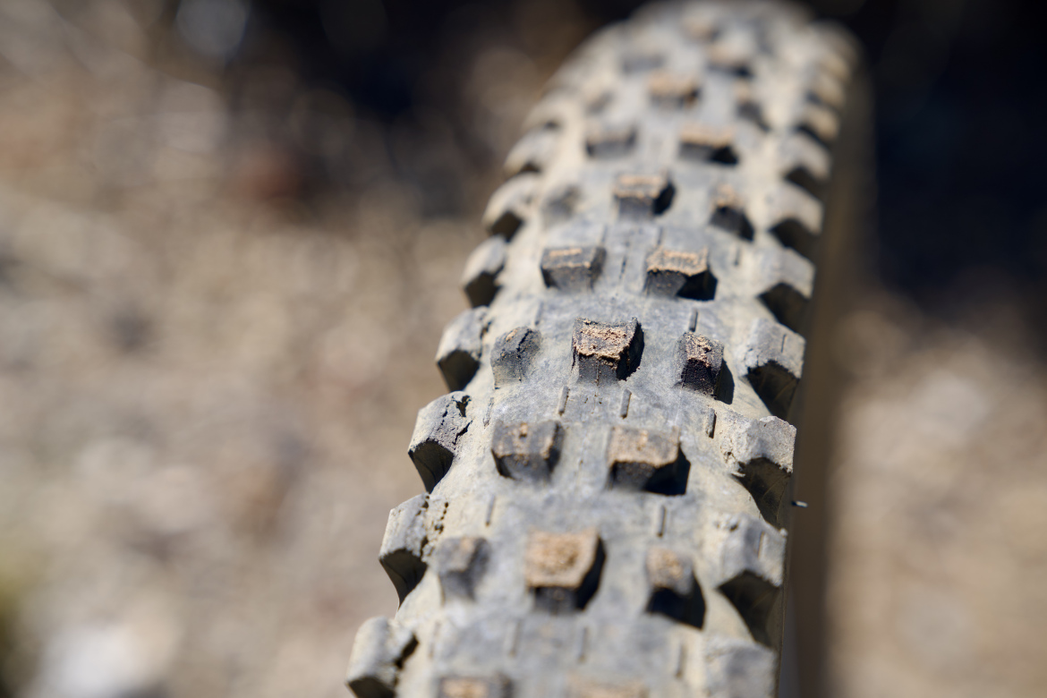 Close-up of a worn mountain bike tire, showing the textured surface and tread patterns with some dirt particles clinging to it. The background is out of focus, emphasizing the tire's details.