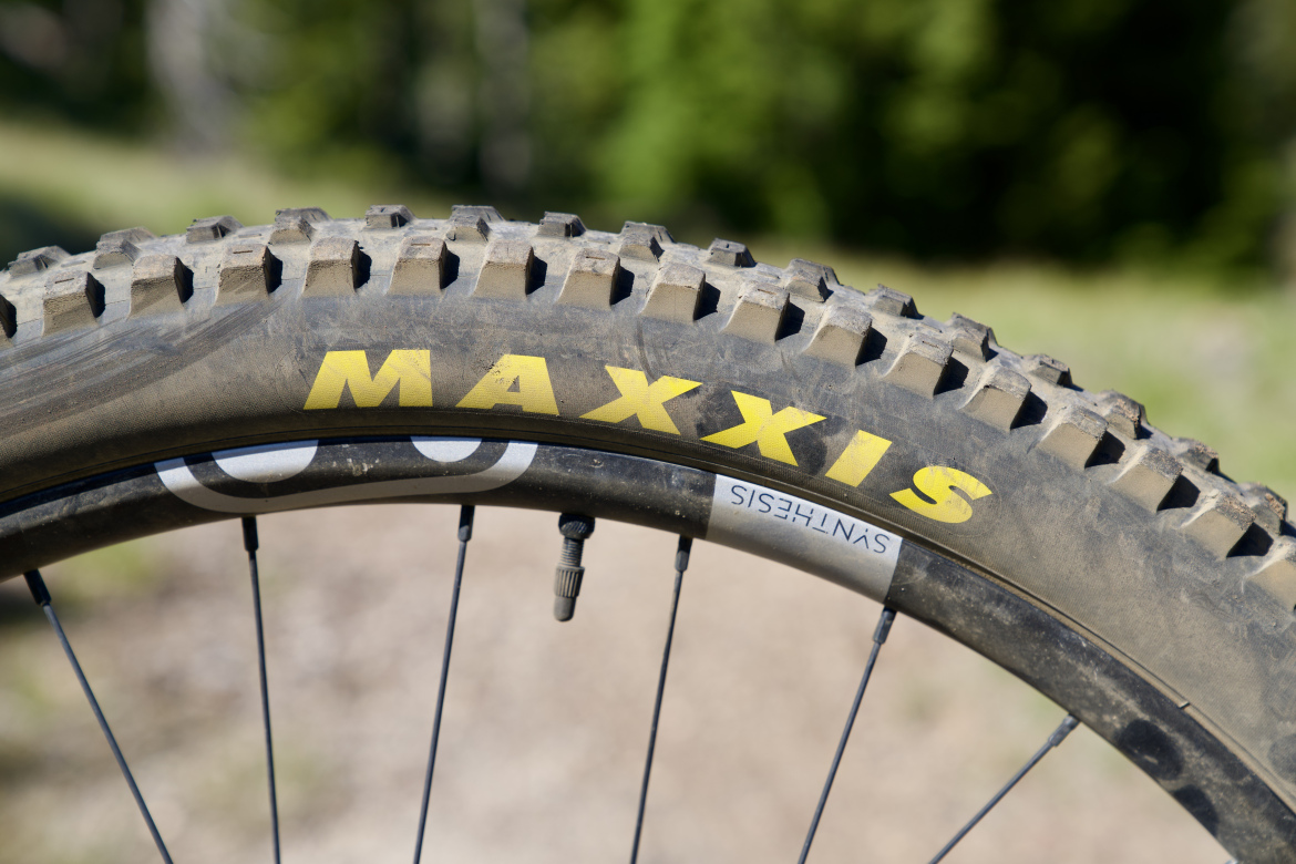 Close-up view of a mountain bike tire, featuring prominent tread patterns and the brand name "MAXXIS" in yellow lettering. The tire is partially dirty, indicating use on outdoor terrains, with a blurred green background suggesting a natural setting. 