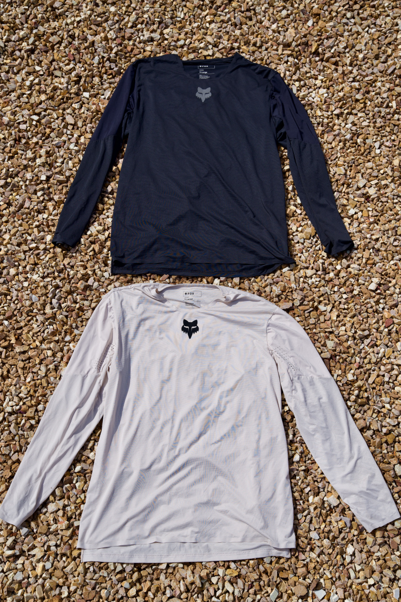 Two long-sleeve shirts are laid out on a gravel surface. The top shirt is navy blue, featuring a logo near the neckline. The bottom shirt is light gray, also with a logo in a similar position. The fabric appears smooth and lightweight, suitable for active wear.