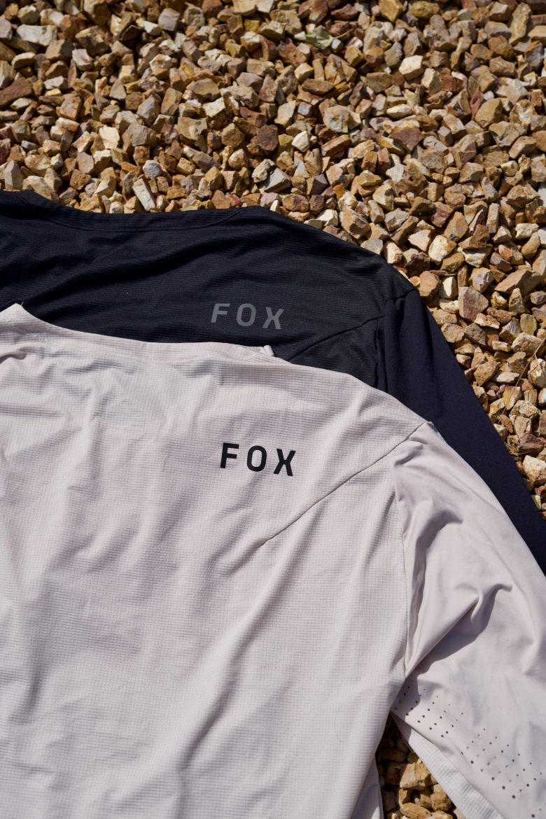 Two athletic jerseys laid flat on a textured surface made of small gravel stones. The jersey on the top is black, while the one below is light gray. Both jerseys feature the word "FOX" printed on the back in bold lettering. The fabric appears lightweight and breathable, suitable for sports or outdoor activities.
