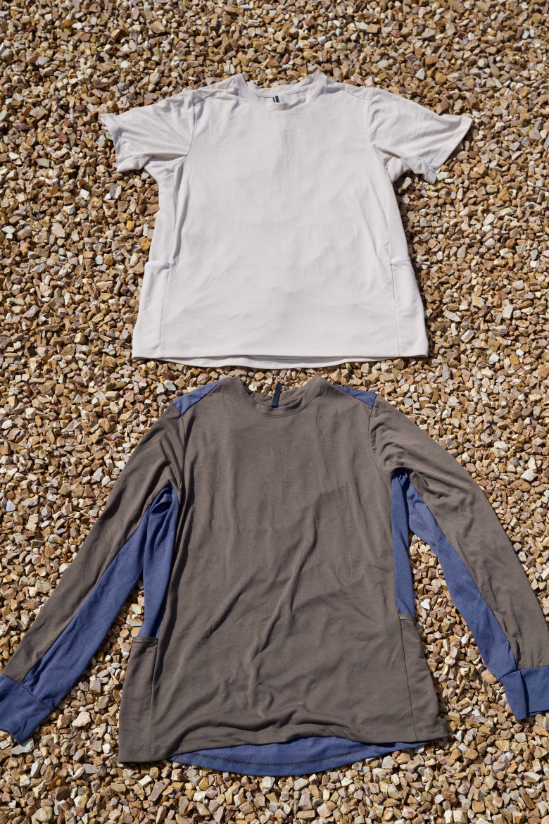 A light gray short-sleeve t-shirt is placed on top of a darker long-sleeve shirt with blue accents, both resting on a bed of small, naturally colored stones.